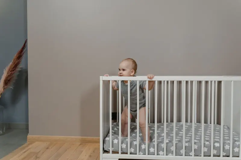 A cute baby in a grey onesie stands in a white crib indoors