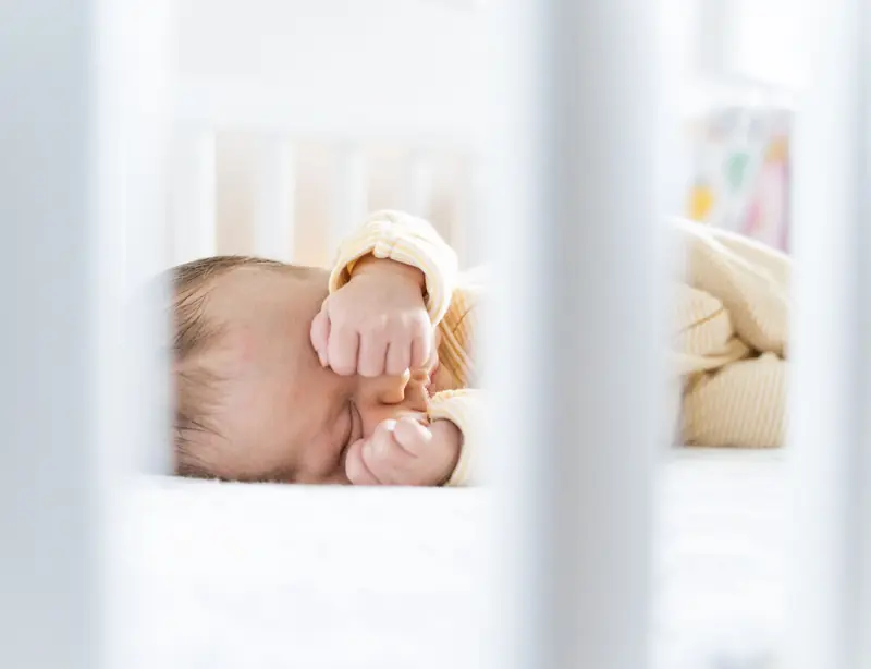 A serene sleeping baby rests peacefully in a bright and cozy crib, framed by white bars